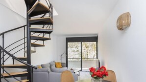 Ein modernes Interieur mit einer Wendeltreppe, einem grauen Sofa, einem runden Tisch mit Blumen und einem Fenster mit Blick auf Bäume.