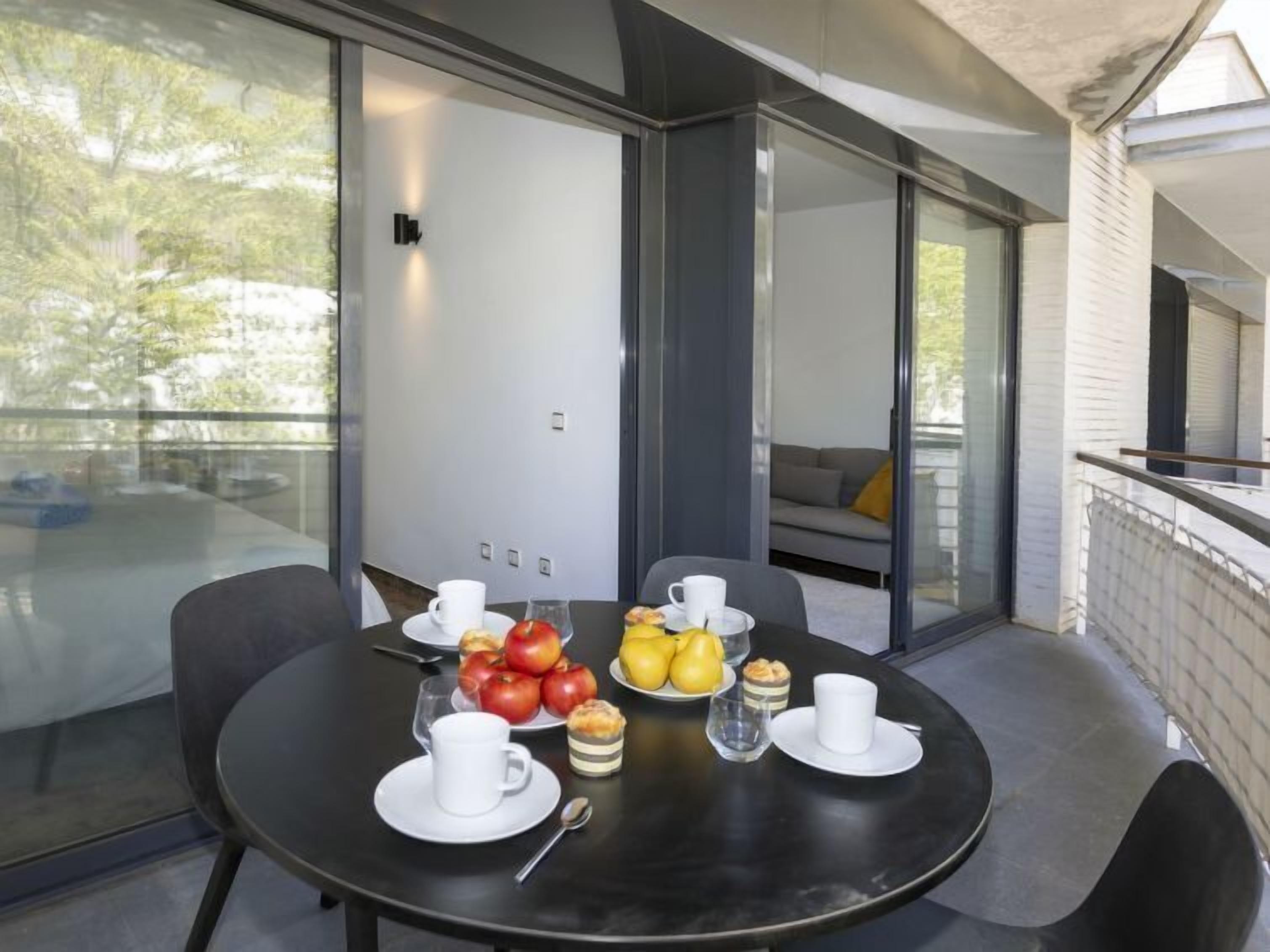 A modern dining area with a round table set for breakfast, featuring fruits, pastries, and white cups.