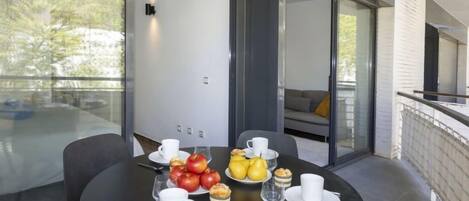 A modern dining area with a round table set for breakfast, featuring fruits, pastries, and white cups.