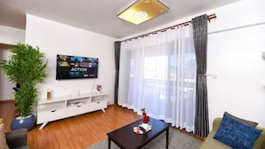 Una sala de estar moderna con una televisión de pantalla plana, una mesa de centro, un sofá, una planta en maceta y un amplio ventanal con cortinas translúcidas.