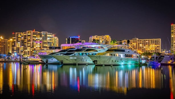 A marina with several yachts docked at night, reflecting city lights on the water.
