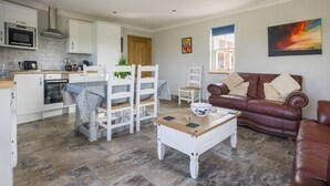 A modern kitchen with white cabinets, a microwave, and a dining area with white chairs. There is a brown leather sofa and a coffee table in the living area. - The Shamba, Bainvalley Cottages (Hemingby)