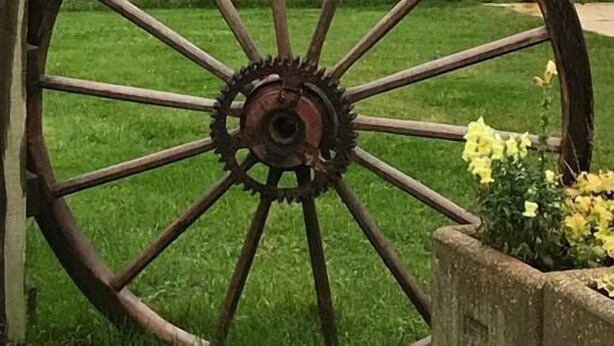 A large wooden wheel with a metal gear in the center, set against a rural landscape with a wooden fence and fields. - The Shamba, Bainvalley Cottages (Hemingby)