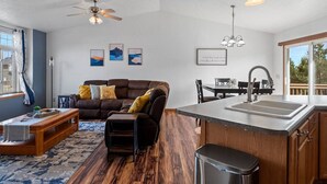 Un salon moderne avec un canapé en cuir marron, une table basse en bois et un tapis. On y trouve également un coin repas avec une table et des chaises, ainsi qu’une cuisine équipée d’un évier et d’un plan de travail.