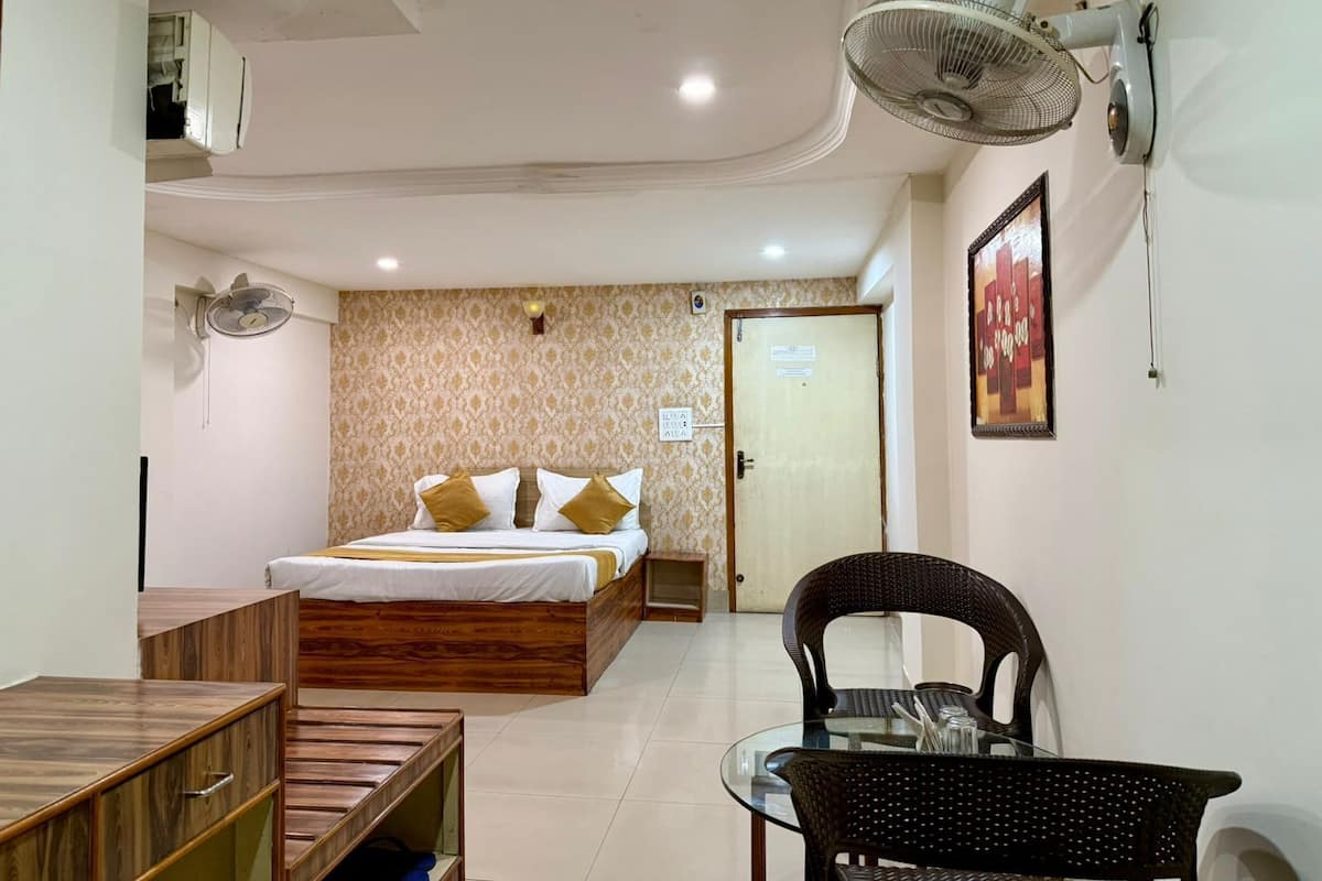 Habitación de hotel con cama, escritorio de madera, mesa de vidrio, silla, ventilador de pared y puerta.