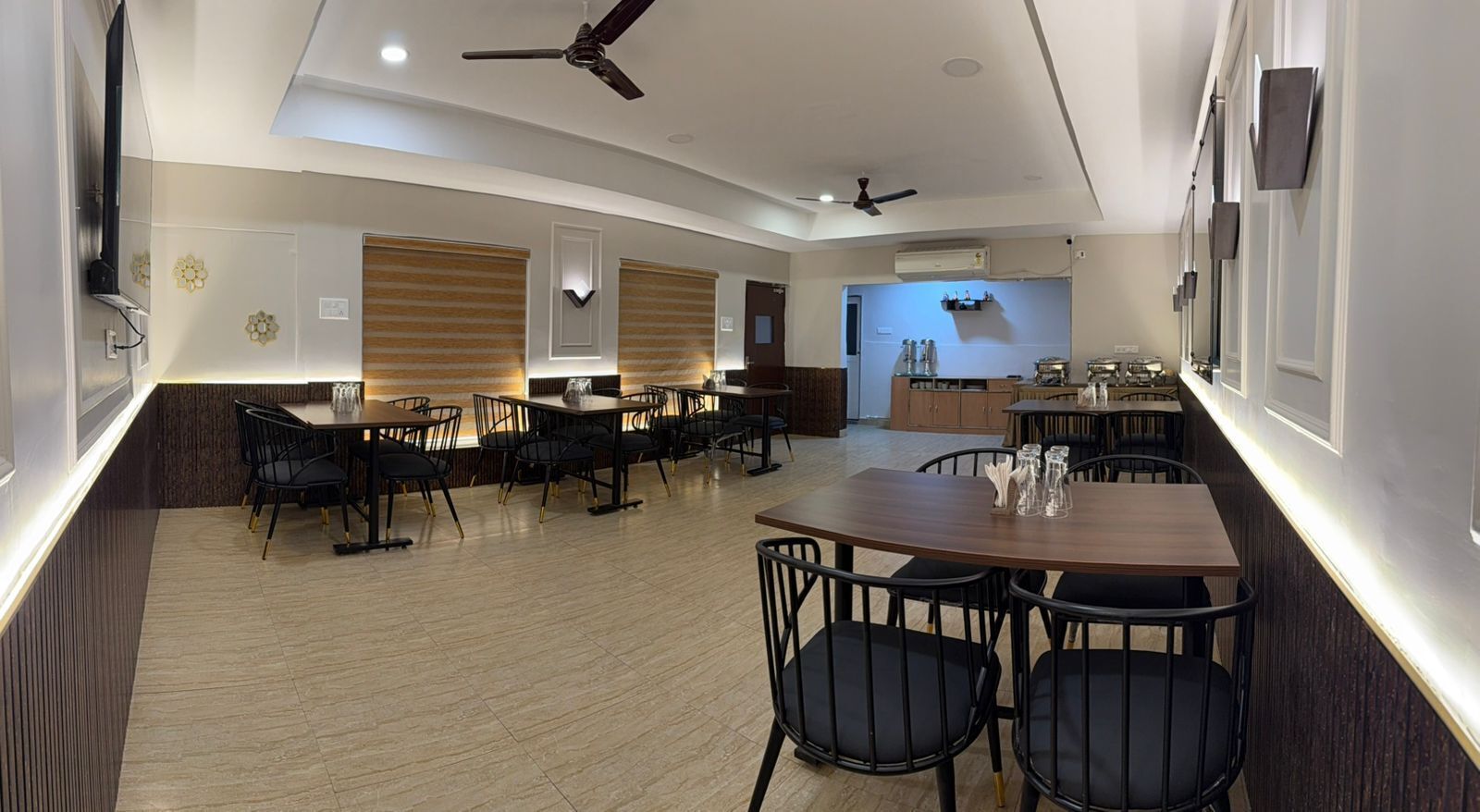 A modern dining area with multiple tables and chairs, ceiling fans, and a kitchen in the background.