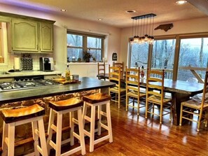 A kitchen with a central island, bar stools, wooden chairs, and large windows offering a view of trees. - Beech Mtn Getaway · Mountain Getaway, ⛳ Golfing, Hiking, Wifi +Netflix (Beech Mountain)