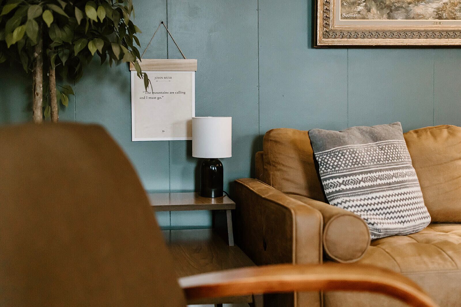En hyggelig stue med en brun lædersofa, et lille sofabord med en lampe og et indrammet citat på væggen.