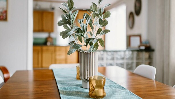Une table à manger ornée d’un vase rempli de feuilles vertes et d’une bougie.