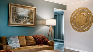Une pièce avec un canapé en cuir marron, un tableau représentant un paysage encadré, une lampe sur pied et une tapisserie murale tissée.