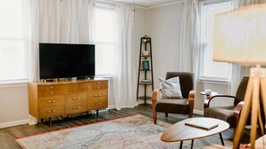 Un salon avec un téléviseur à écran plat posé sur une commode en bois, un fauteuil marron, un tapis à motifs et une petite table ronde sur laquelle se trouve un livre.