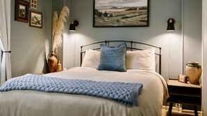 Une chambre à coucher comprenant un lit, deux tables de chevet, une petite table, une lampe fixée au mur, un tableau représentant un paysage encadré et un vase contenant des plantes séchées.