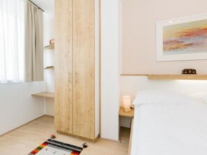 A bedroom with a bed, a wooden wardrobe, a bedside table with a lamp, a shelf with a clock, and a painting on the wall.