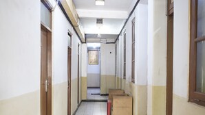 A long, narrow hallway with white tiled floors and beige walls. There are several doors on the left and right, and a few benches along the right side. - SPOT ON 91444 Hotel Arimbi (Bandung)