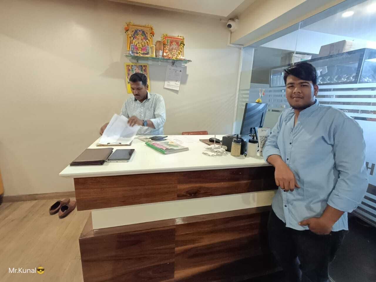 Un mostrador de recepción con acabado de madera y superficie blanca. Dos personas están detrás del mostrador, una de ellas parece ser la recepcionista.