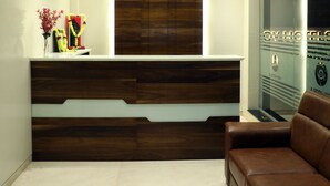 A reception area with a wooden reception desk, a brown leather couch, and a glass wall with a logo. - VOV Hotels Vijayapura (Bijapur)