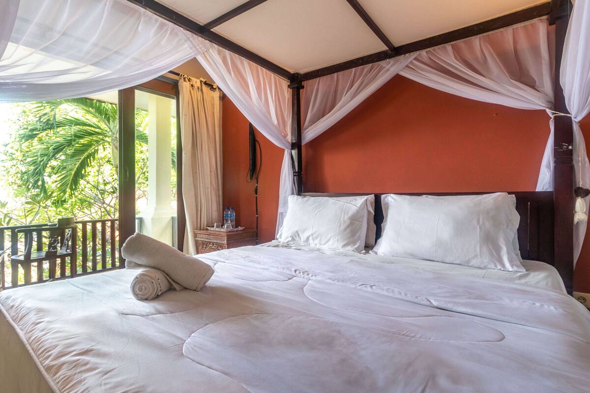 Una cama con sábanas blancas, dosel y vista a palmeras.