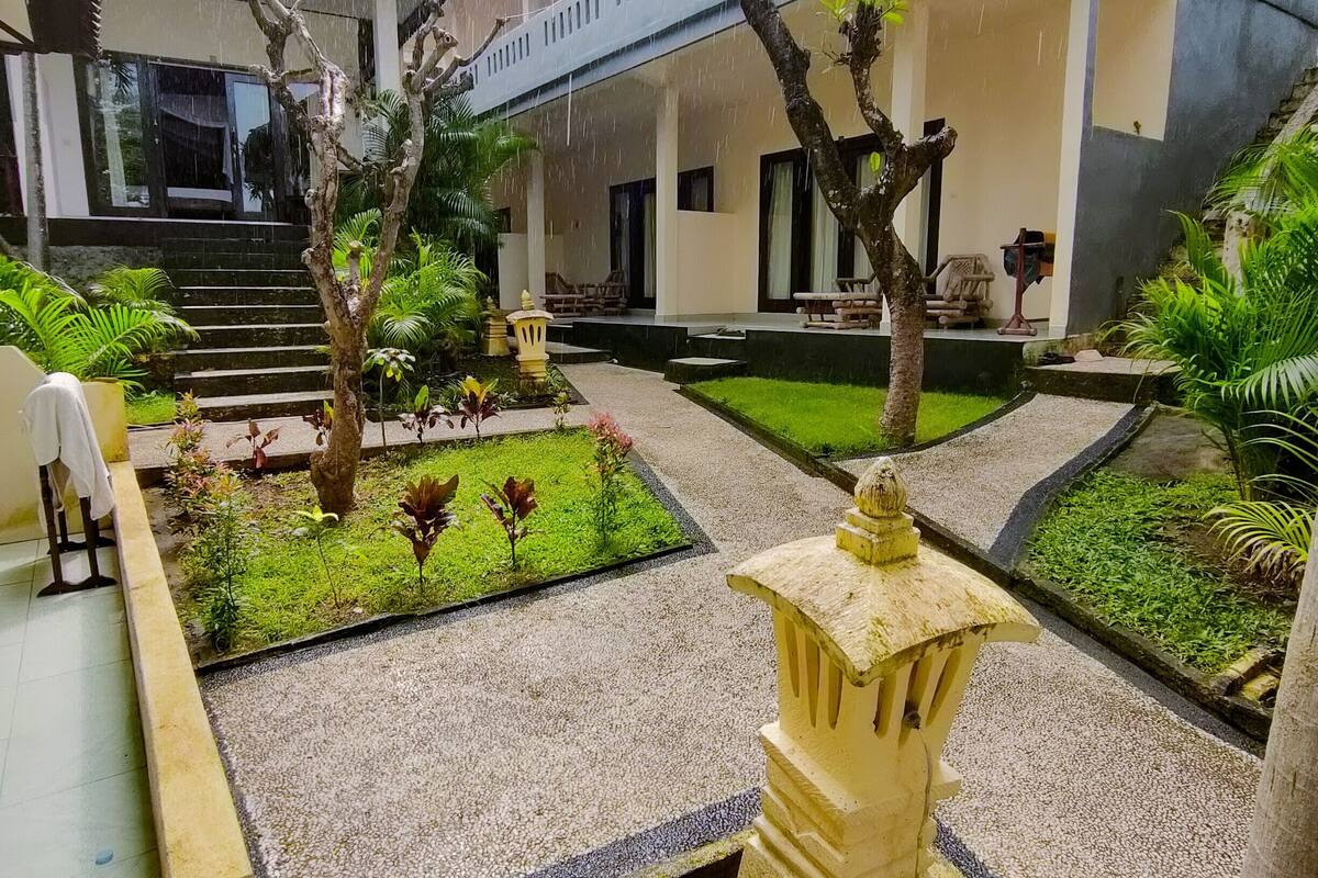 Un patio con un sendero de piedra, un pequeño jardín con diversas plantas, un farol de piedra decorativo y un edificio con techo de tejas y ventanas grandes.