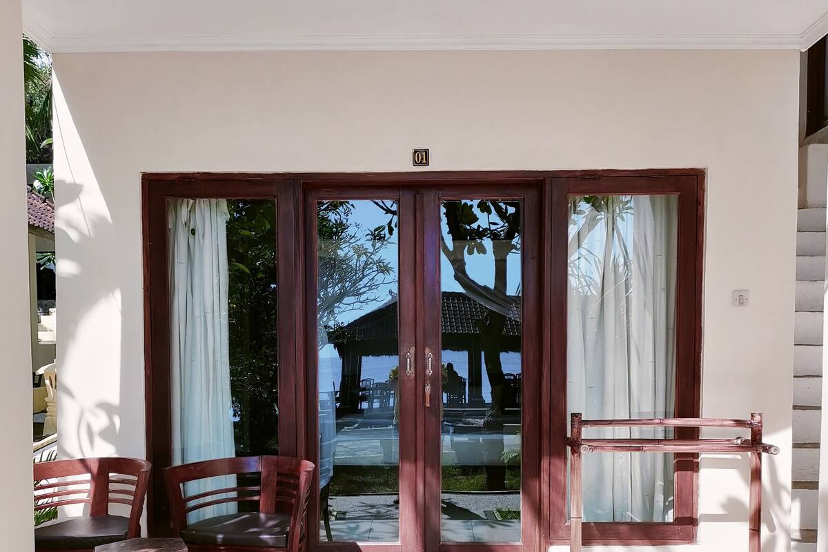 Una habitación con puerta corrediza de vidrio que da a vista de playa y un pabellón. Hay dos sillas de madera y una pequeña mesa con un adorno.