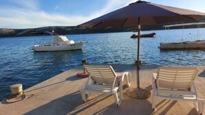 A beachside area with two white lounge chairs, a large umbrella, and a boat in the background. - House in a quiet environment in the immediate vicinity  of the beach (Stara Novalja)