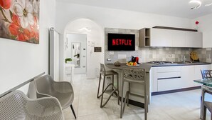 Una cucina moderna con un'ampia televisione a schermo piatto che mostra 'NETFLIX', un'area pranzo con sedie e un'isola cucina con piano cottura a gas.