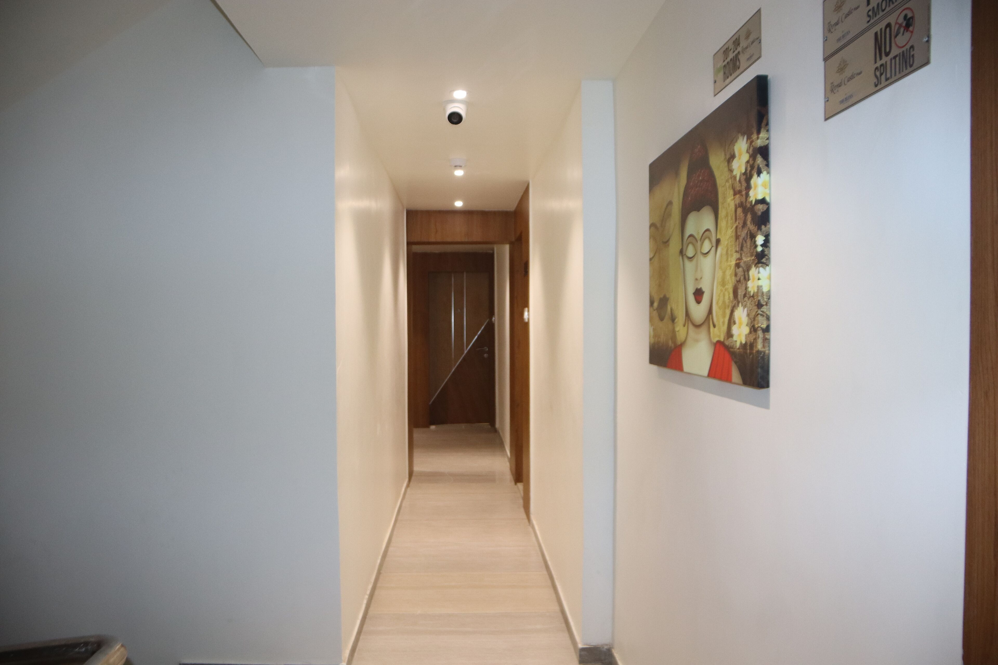 Un couloir avec une porte en bois au fond, une caméra de surveillance fixée au plafond et un panneau mural.