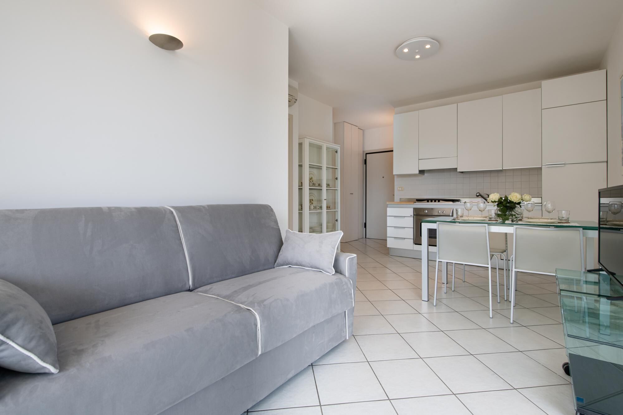 Una sala de estar moderna con un sofá gris, una mesa de comedor de vidrio y armarios blancos al fondo.
