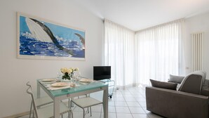 A modern living room with a glass dining table set for four, a grey sofa, a flat-screen TV, and a large abstract painting on the wall. - Gran Torino 17 in Cattolica (Cattolica)