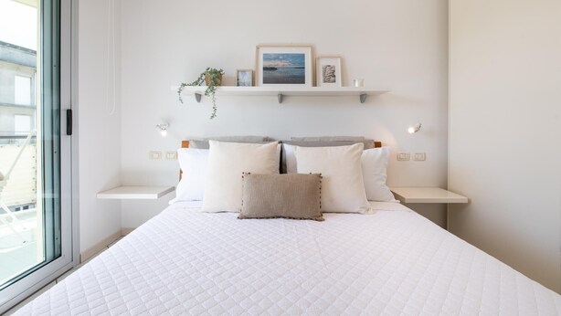A bedroom with a large bed, white walls, and a window. There are two bedside tables with lamps, a shelf above the bed with decorative items, and a view of the outside through the window. - Gran Torino 17 in Cattolica (Cattolica)