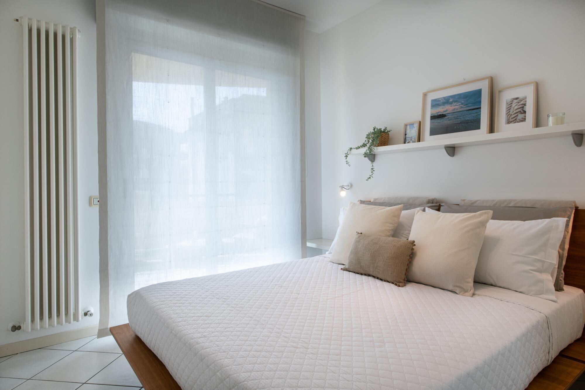 A bedroom with a large bed, a radiator, a window with sheer curtains, and a shelf with framed pictures and a plant.