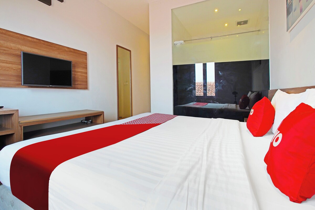 A hotel room with a bed, a TV mounted on the wall, a wooden desk, and a large mirror.
