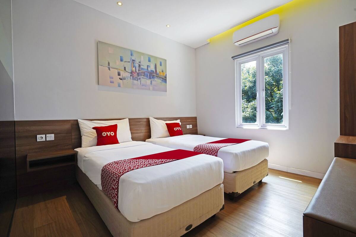 A hotel room with two beds, a painting on the wall, a window with a view of trees, and an air conditioning unit.
