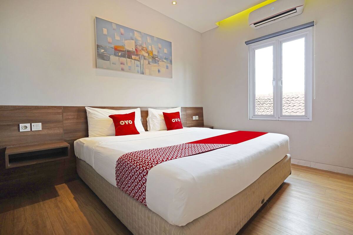 A modern hotel room with a large bed, a red and white patterned throw, and a cityscape painting on the wall.