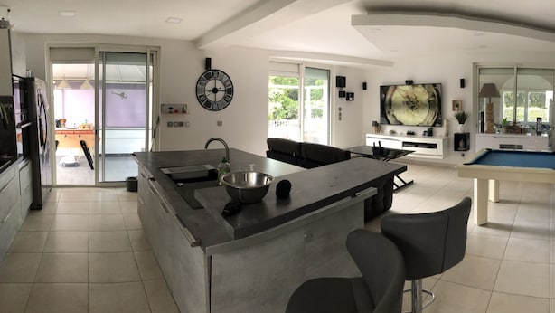 Una cocina moderna con una isla central, un comedor con mesa de billar y una sala de estar con televisor grande.