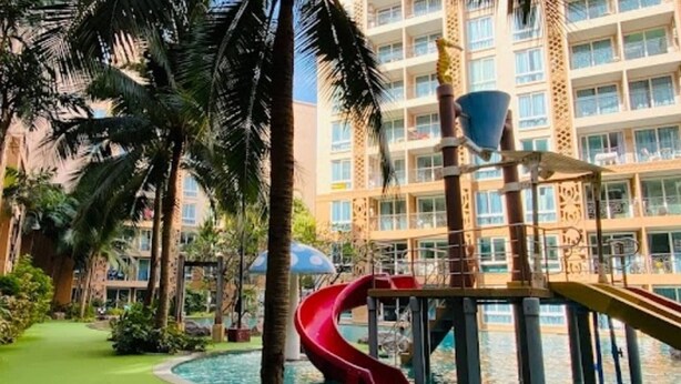 Zona de piscina con un tobogán rojo, palmeras y un edificio con balcones al fondo.