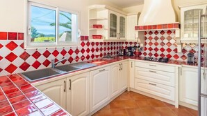 Een keuken met witte kastjes, een spatwand in rood en wit, een raam met uitzicht op palmbomen, en een fornuis.