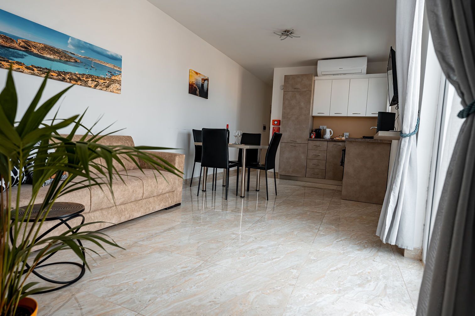 A modern living room with a beige sofa, a dining area with a table and chairs, and a kitchen area with cabinets and a countertop.