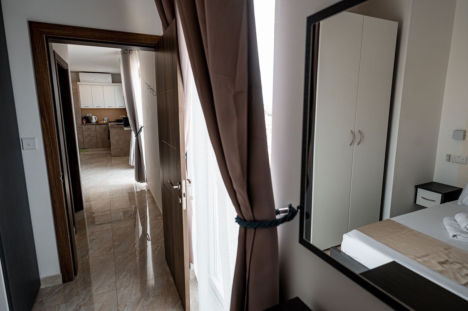Habitación de hotel con cama, mesita de noche, espejo y vistas de la cocina y el área de estar a través de una puerta abierta.