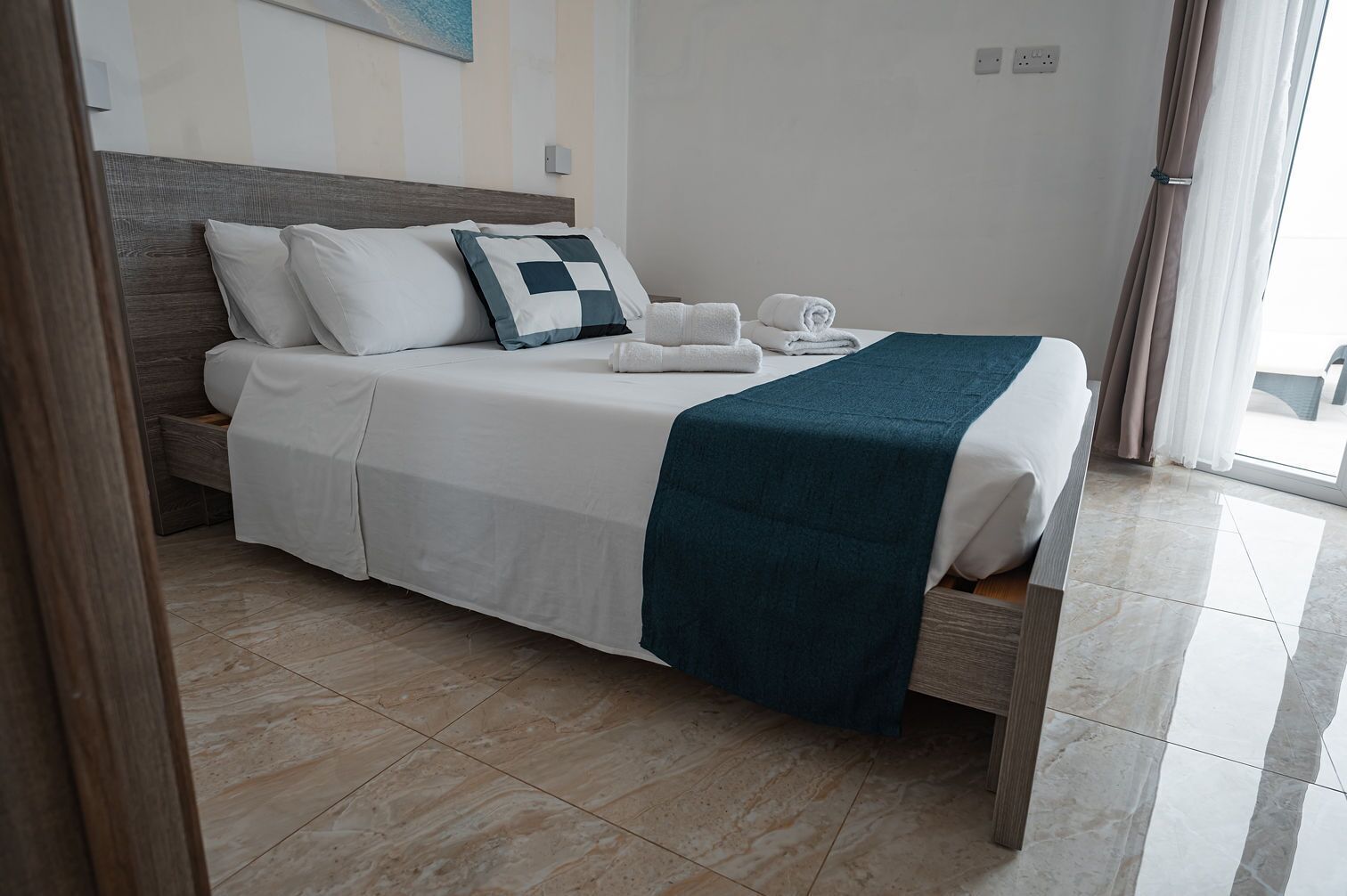 A neatly made bed with white bedding and a teal throw blanket, a wooden headboard, and a tiled floor.