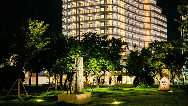A multi-story hotel with a curved facade, illuminated signage, and a well-maintained lawn with trees and a sculpture. - Jackson9s Hotel (Chuncheon)