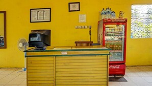 A yellow wall with a red Coca-Cola refrigerator, a black television, and a wooden counter labeled "KASIR".