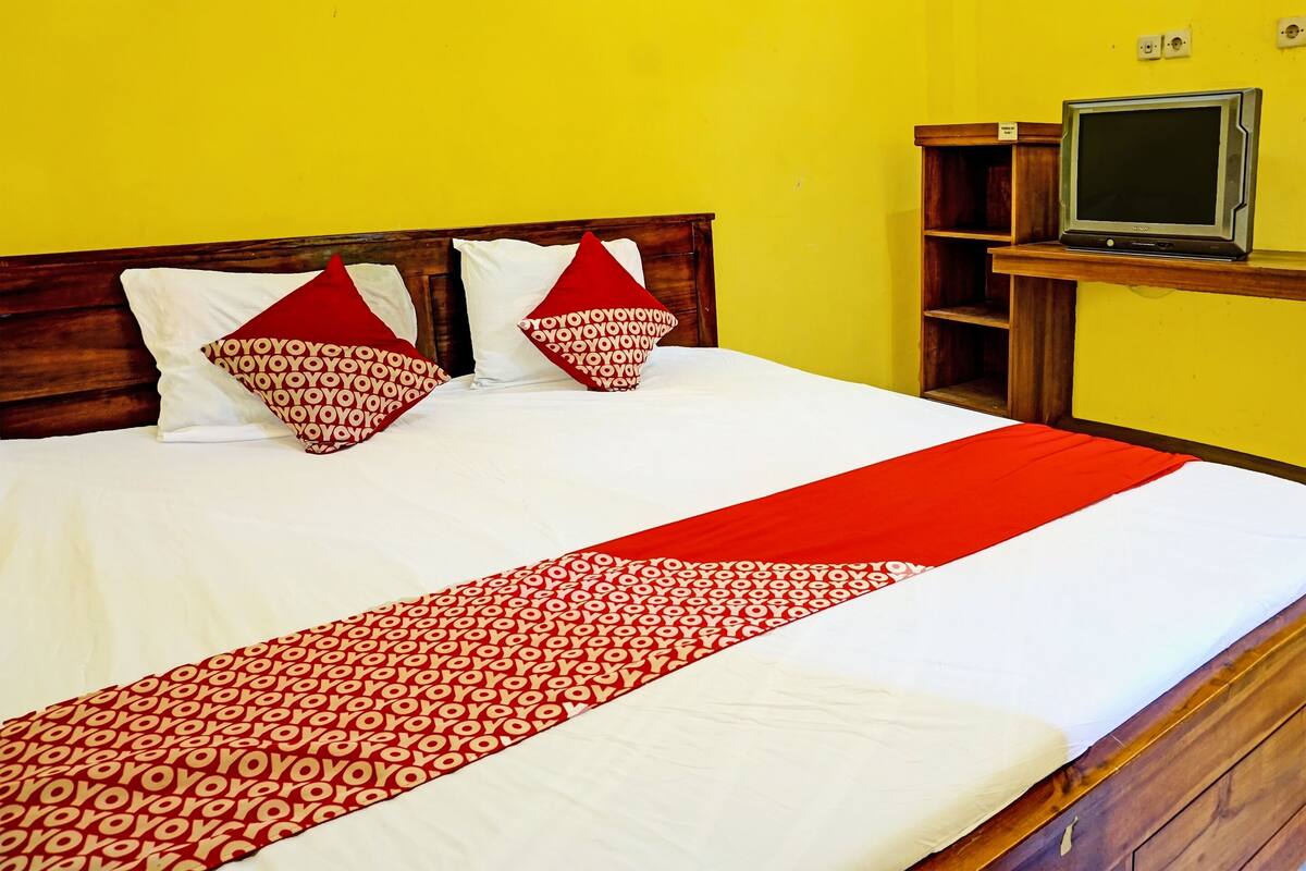 Una cama bien hecha con una colcha roja y blanca de estampado, dos almohadas rojas con un diseño circular, y un televisor sobre un mueble de madera frente a una pared amarilla.
