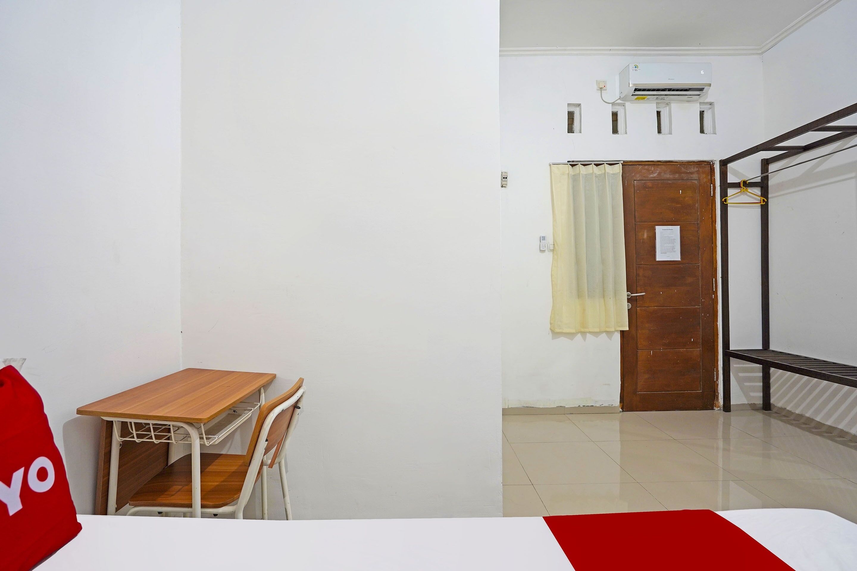 Una habitación con un escritorio de madera, una silla, una cama con edredón rojo y blanco, una puerta de madera y un banco de metal.