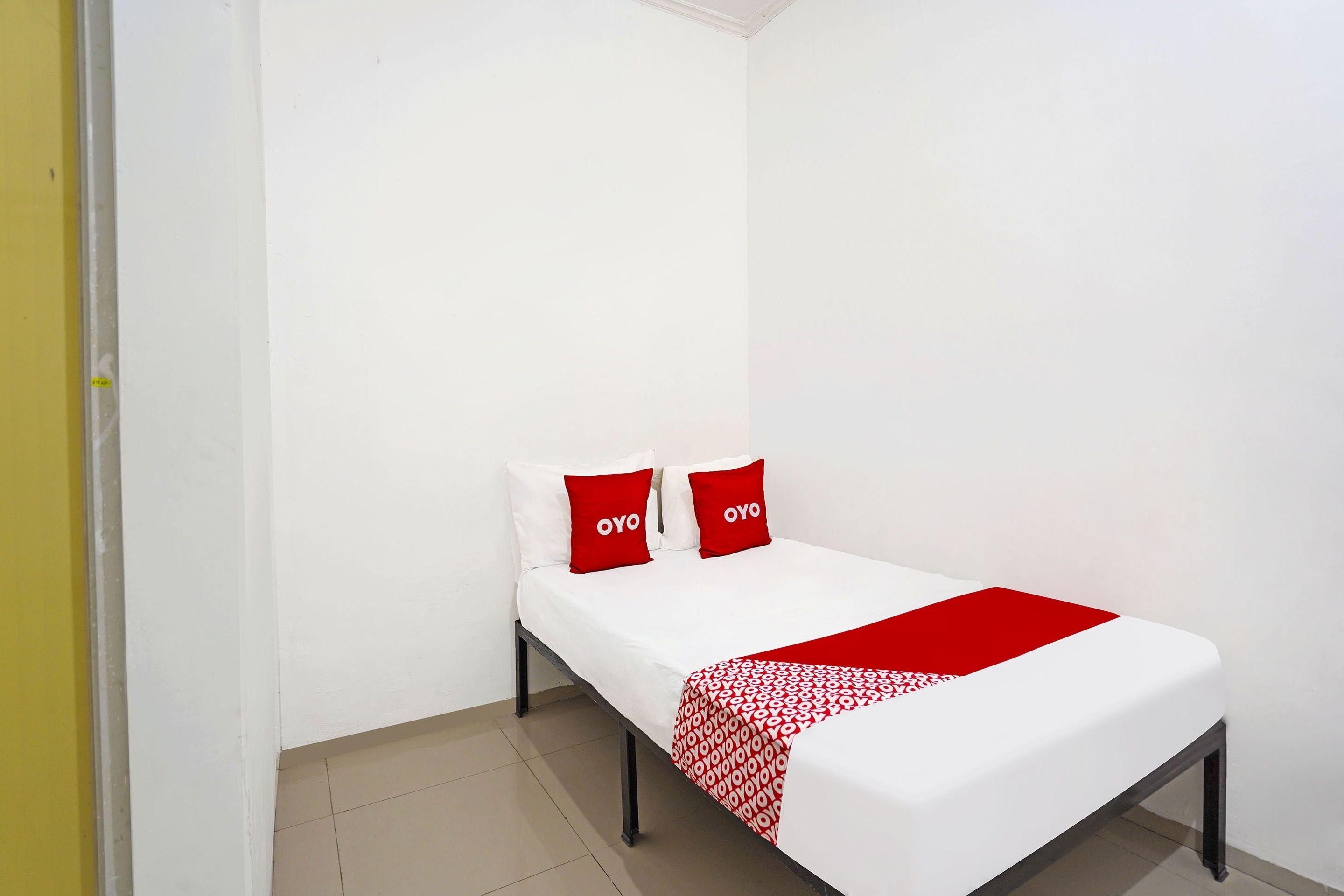 A small, single-bed room with a white bedspread, two red pillows, and a red and white patterned throw blanket.