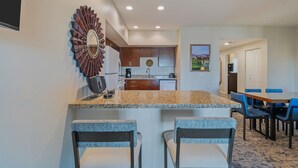 A modern kitchen with granite countertops, wooden cabinets, and a built-in oven. There is a dining area with blue chairs and a table, and a kitchen island with two bar stools. - Coachella Shuttle, Lazy River: 3BR in Resort (Indio)