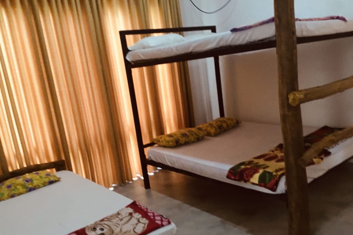 A bunk bed with a white mattress and a floral-patterned blanket. There is a chair with a yellow cushion and a table with a white tablecloth and a teddy bear decoration.