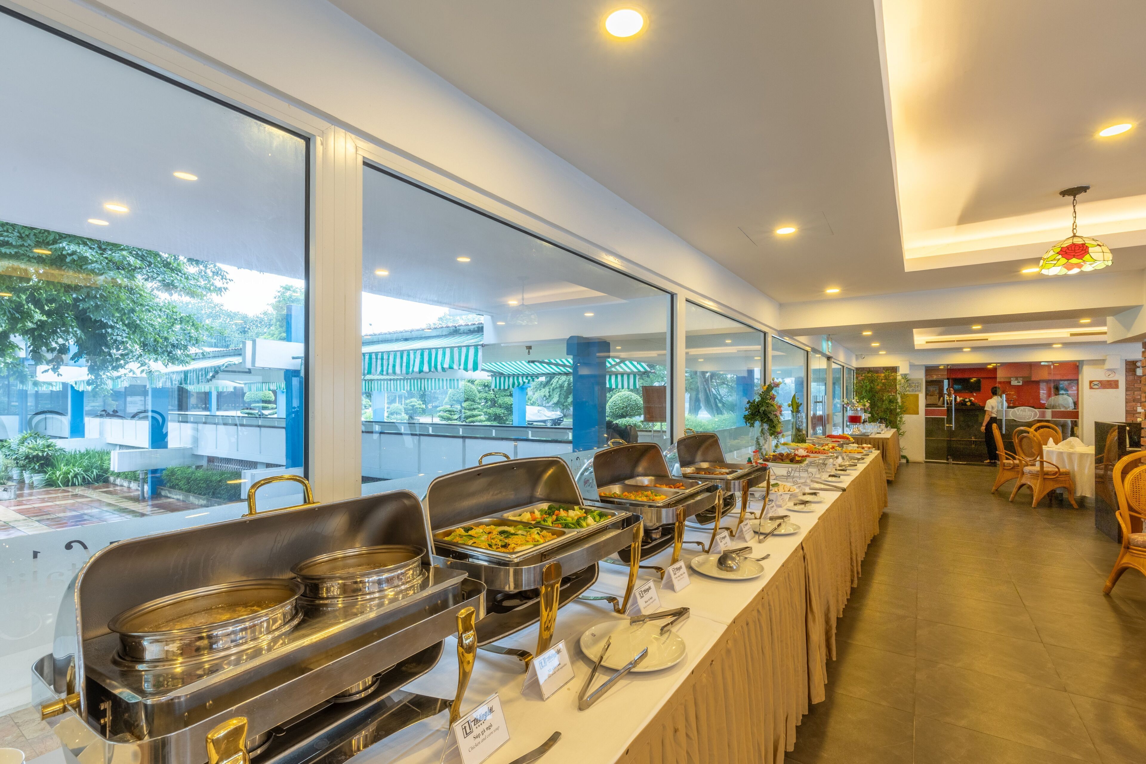 A buffet setup with multiple serving stations, a long table with dining chairs, and a view of the outdoors through large windows.