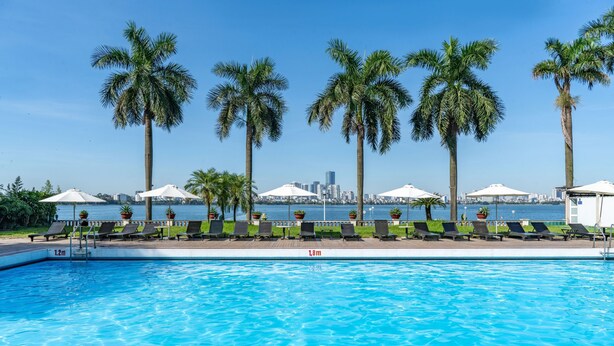 Ein Swimmingpool mit Liegestühlen und Sonnenschirmen, Palmen und einer Stadtkulisse im Hintergrund.