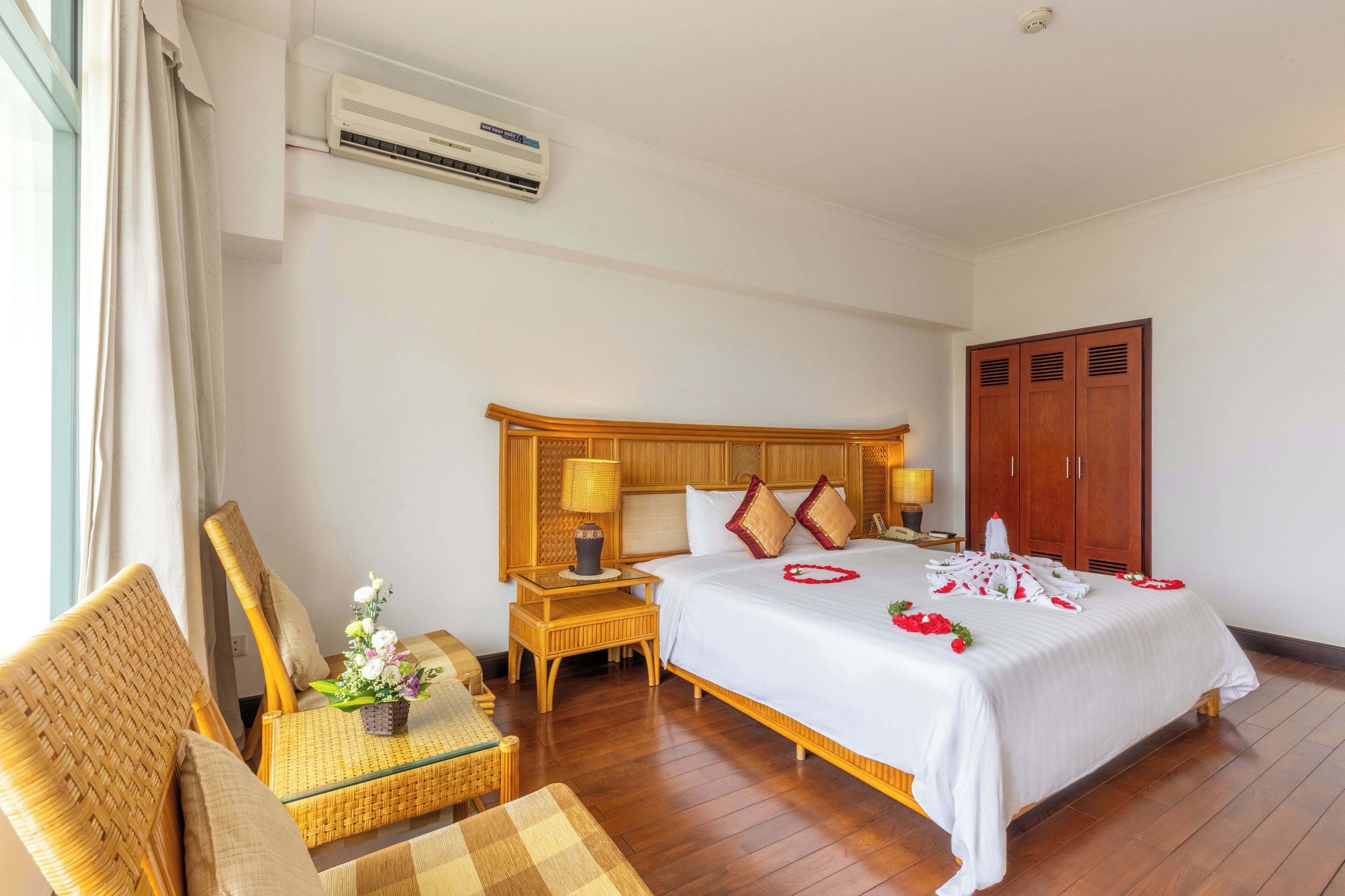 A hotel room with a wooden bed, a nightstand with a lamp, a wicker chair, and a small table with a vase of flowers.