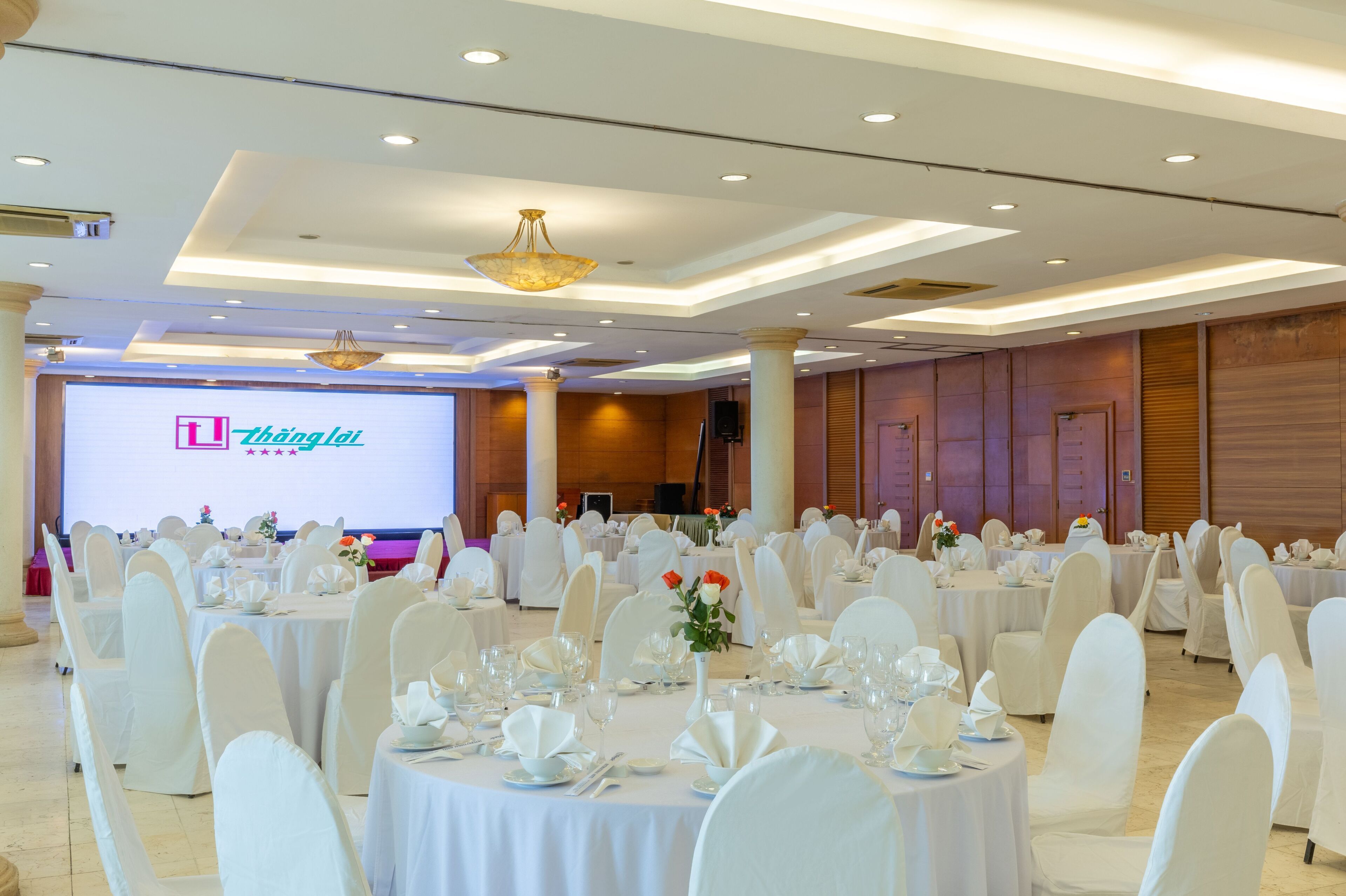 A banquet hall with round tables set for events, white chairs, and a large screen displaying a presentation.